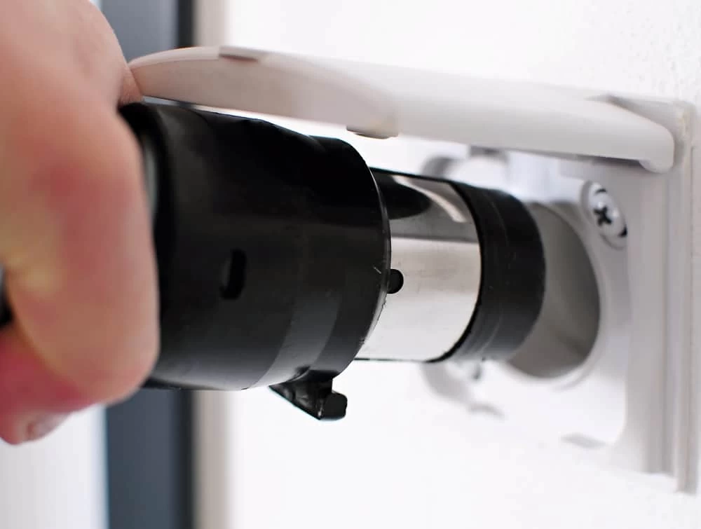 Close-up of a hand using a pcentral vacuum LIower drill to install or remove a screw in a white electrical socket mounted on the wall, capturing the precision essential for tasks, like setting up central vacuum systems in Long Island, NY. The focus is on the interaction between the drill and socket.