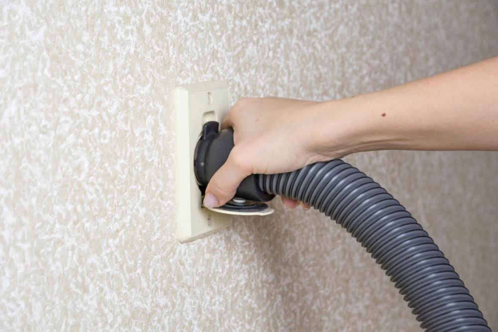 A hand plugs a vacuum hose into a wall outlet, set against a textured, light-colored wall. The outlet is expertly designed for central vacuum systems in Long Island NY, offering convenience and efficiency.