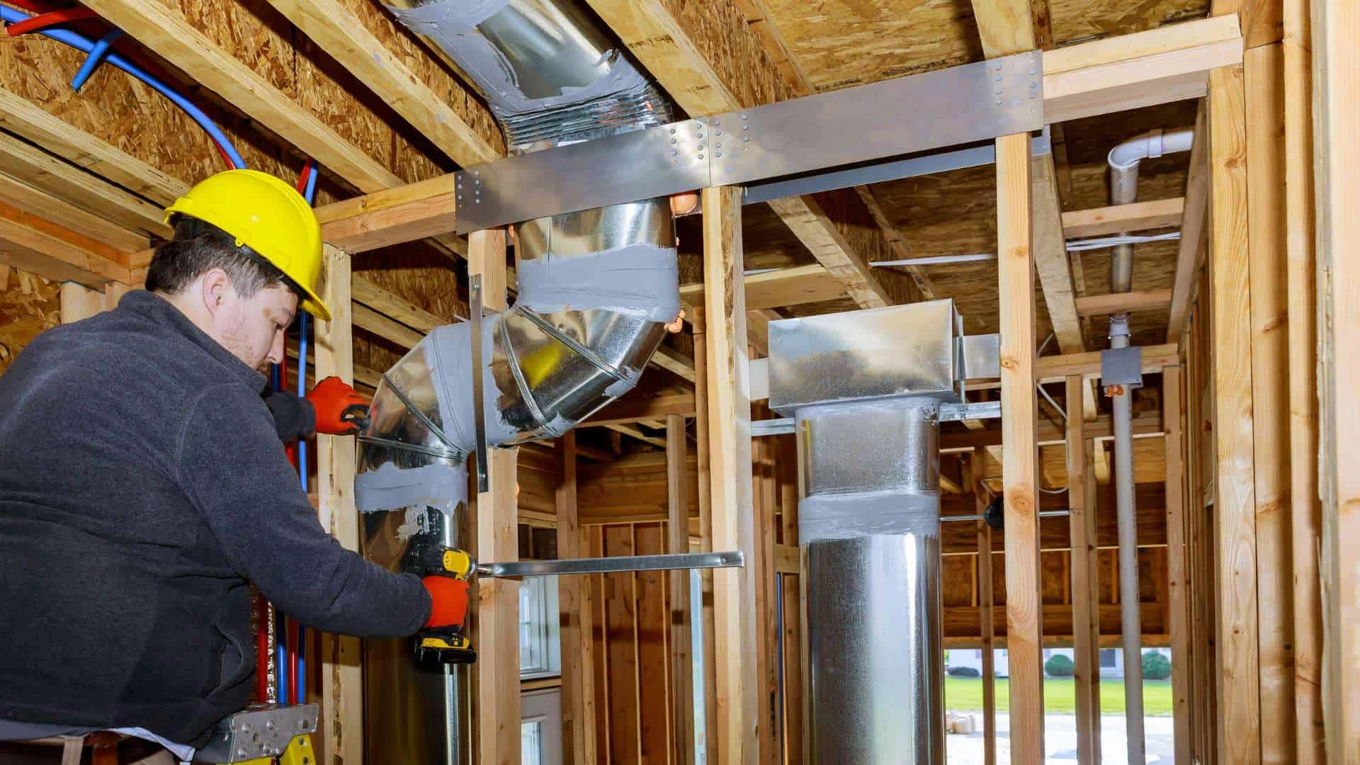 A construction worker in a yellow hard hat and orange gloves is using a power drill to install ductwork for central vacuum systems within the wooden framework of a building on Long Island, NY. Several ducts and pipes are visible, and the interior is unfinished with exposed beams.