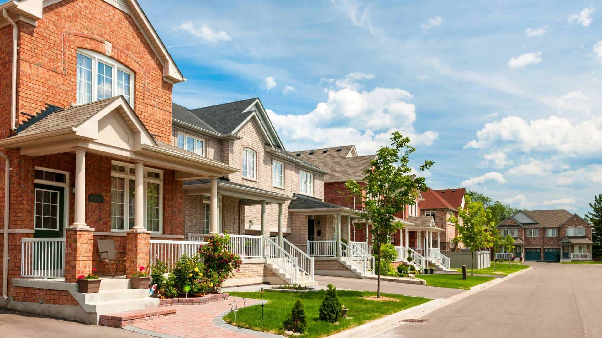 A row of suburban houses with brick and siding exteriors, front porches, and well-maintained lawns line a quiet, sunny street in Long Island, NY. Beneath the clear blue sky and scattered clouds, some homes boast modern features like central vacuum systems.