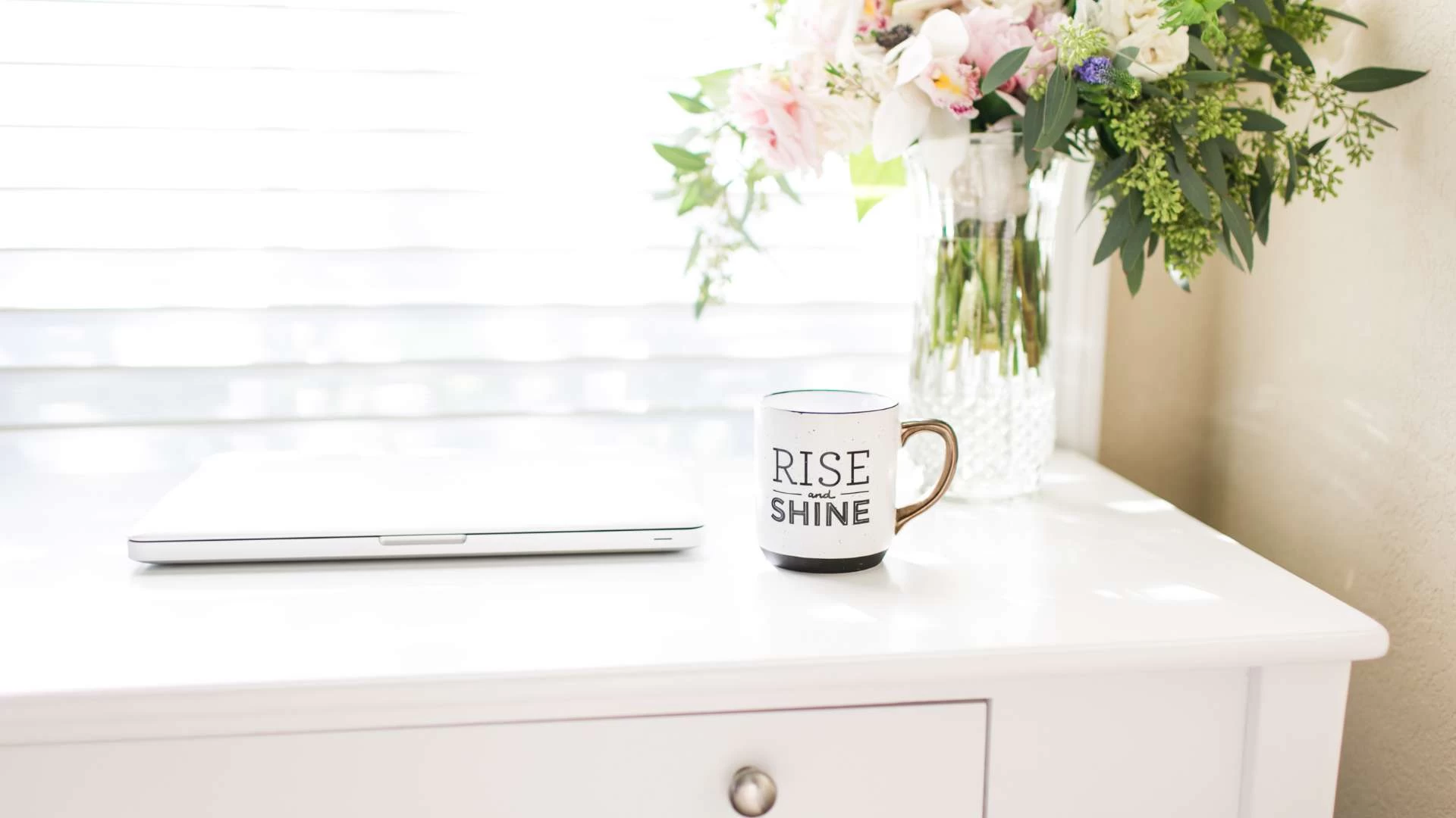 A white table with a closed laptop and a coffee mug that reads "Rise & Shine" sits next to a vase of colorful flowers. Sunlight filters through the window, creating a bright atmosphere reminiscent of an ideal Long Island morning, where central vacuum systems keep everything spotless.