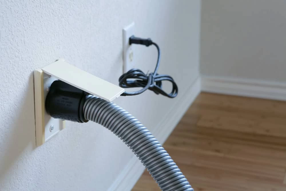 A close-up of a wall features a central vacuum hose connected to an outlet, typical in homes across Long Island, NY. An electrical cord is plugged into a separate socket above, with the cord loosely coiled and hanging against the wall. The floor is finished in light-colored wood.