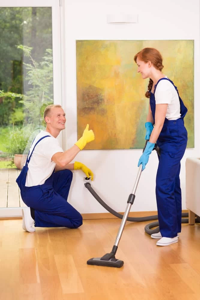 Two people in blue coveralls and gloves diligently clean a room. One expertly wields a vacuum, courtesy of central vacuum systems Long Island NY, across the wooden floor while the other kneels with a thumbs-up. A large abstract painting graces the wall, and greenery is visible through the window.