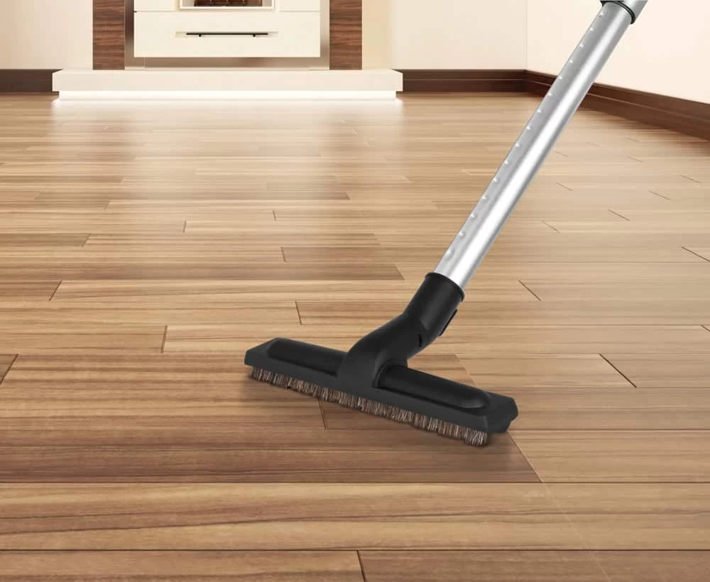 A vacuum cleaner with a brush head glides effortlessly over the light brown, polished wooden floor, showcasing its natural grain. In the background, a cozy fireplace flickers warmly. Experience seamless cleaning with central vacuum systems on Long Island, NY.