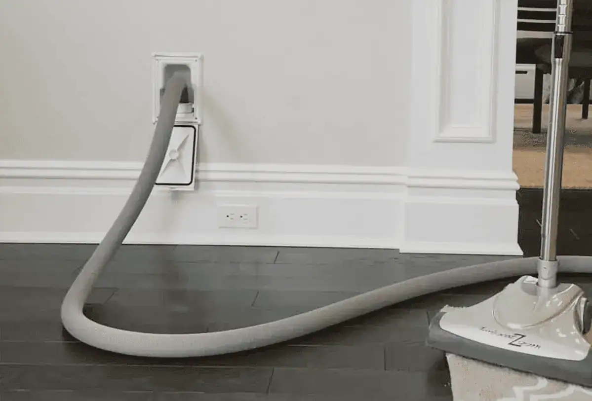 A central vacuum hose plugged into a wall outlet on a white wall, extending across a dark wood floor and connected to a vacuum head resting on a gray and white rug.