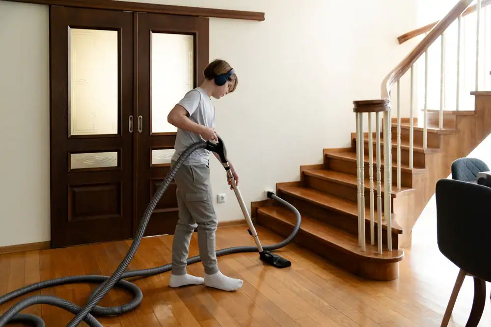 In a bright NY room with wooden accents and double doors, a person wearing headphones vacuums a wooden floor next to a staircase, showcasing the convenience of central vacuum systems Long Island residents love.