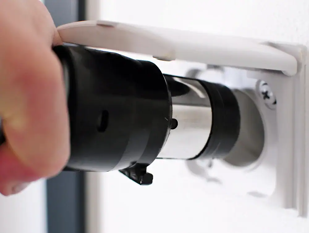 A close-up of a hand plugging a heavy-duty black electrical plug into a white wall socket with a protective cover, commonly seen in homes equipped with central vacuum systems Long Island, NY.