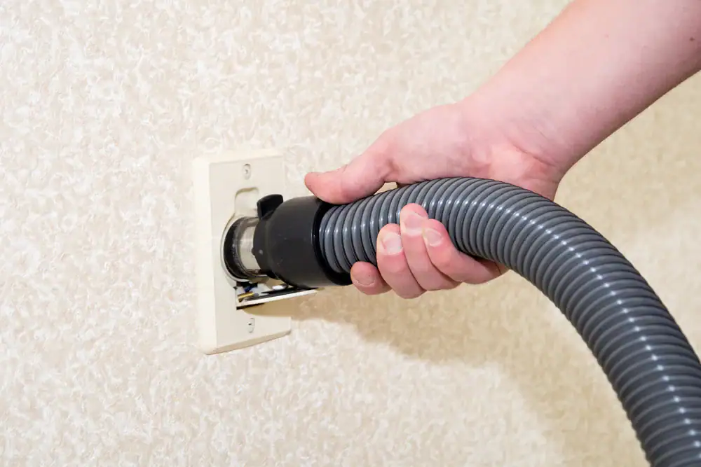 A person’s hand is plugging a vacuum hose into a wall-mounted central vacuum system outlet on a textured, light-colored wall—showcasing the convenience of central vacuum systems in Long Island, NY homes.