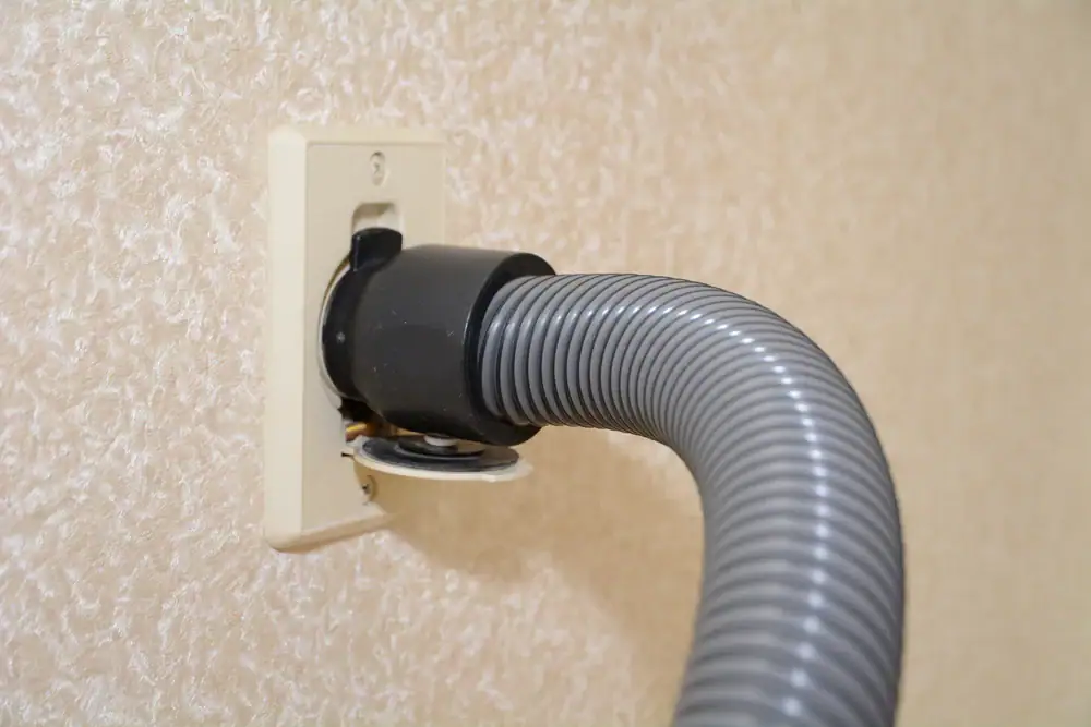A close-up of a gray flexible vacuum hose connected to a wall outlet on a textured beige wall, part of built-in central vacuum systems in Long Island, NY.