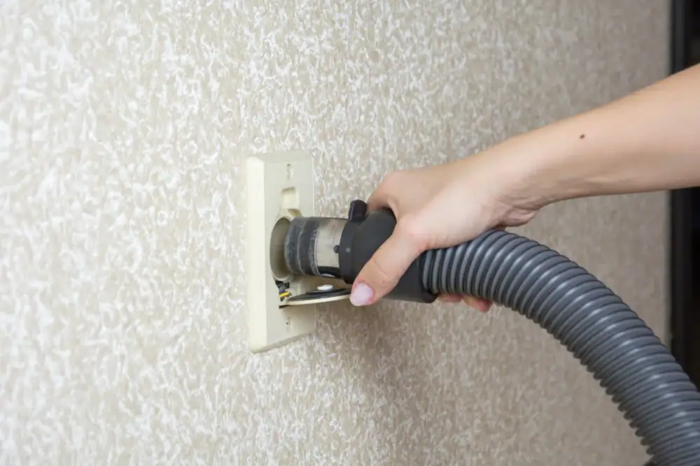A hand inserts a vacuum hose into a wall-mounted central vacuum system outlet on a textured beige wall, showcasing convenient cleaning solutions from central vacuum systems Long Island, NY.