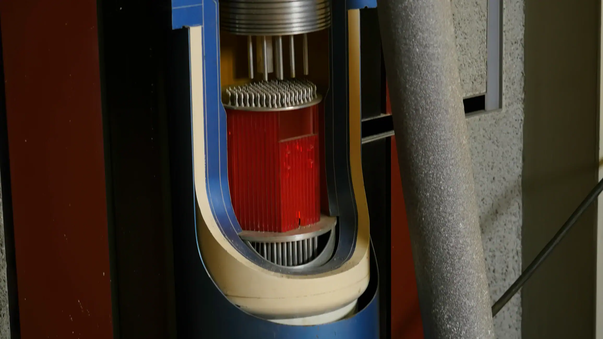 A close-up of a cutaway model of a nuclear reactor, showing internal components like fuel rods and cooling fins, with a red core and blue outer casing. A gray pipe is visible on the right side.