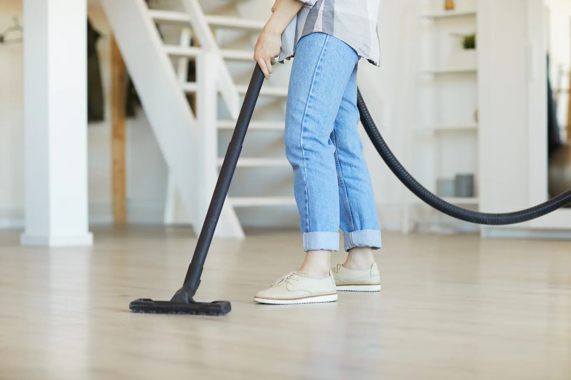 A person wearing rolled-up jeans and beige shoes is vacuuming a wooden floor in a bright, modern NY home with stairs and shelves, using a Central Vacuum System Long Island residents trust.