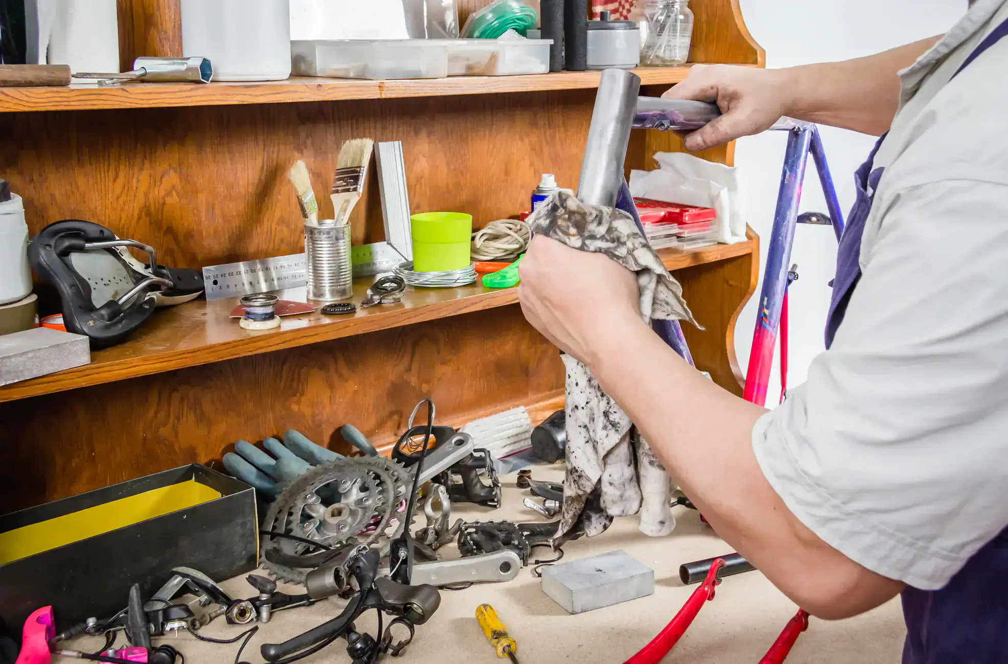 A person cleans a metal bike part with a cloth at a cluttered workbench covered in bicycle tools and components, while shelves hold various supplies in the background, highlighting the need for a Central Vacuum System Long Island, NY.
