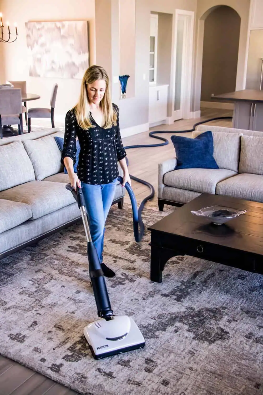 A woman vacuums a rug in a living room with a Central Vacuum System Long Island, NY. She stands between two sofas, holding the vacuum hose, with a coffee table and stylish home decor in the background.