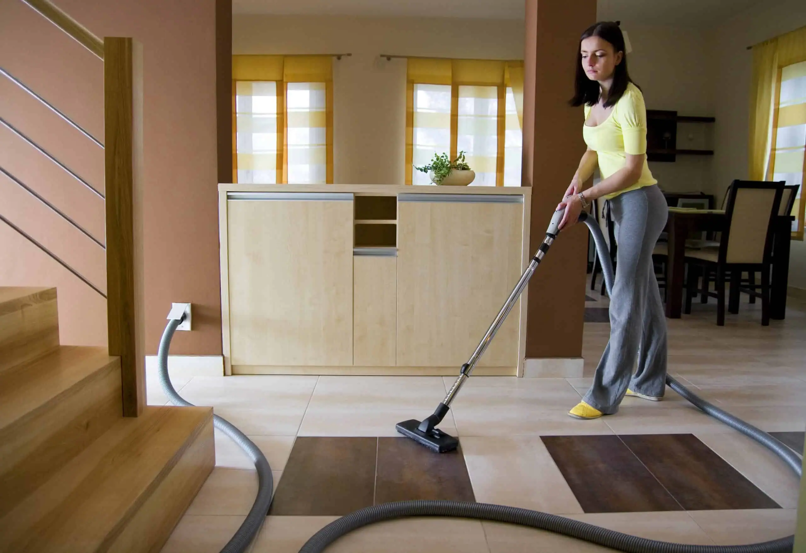 A woman in casual clothes uses a Central Vacuum System Long Island, NY to clean the modern, tidy home with wooden stairs, light tiled flooring, and yellow curtains in the background.