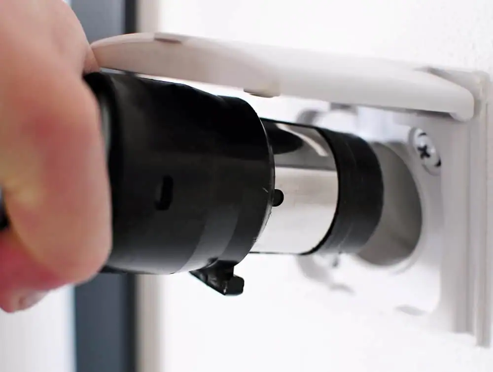 A close-up of a hand plugging a black electrical device into a protective wall socket with a white cover, commonly found in homes equipped with a Central Vacuum System in Long Island, NY.