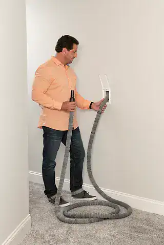 A man in a peach shirt and jeans pulls a long, flexible hose from a Central Vacuum System in a carpeted Long Island, NY hallway.